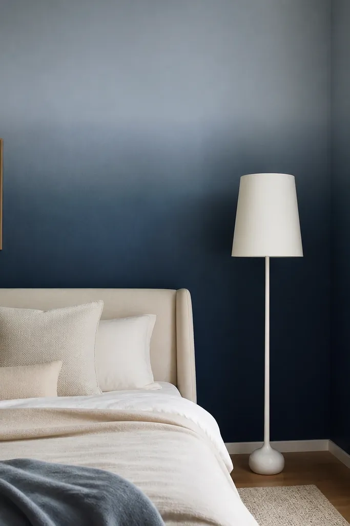 A bedroom wall with an ombre effect: deep navy at the bottom fading to a softer blue-gray near the top. The bed is light beige with pillows in cream. A tall white floor lamp stands near the corner.