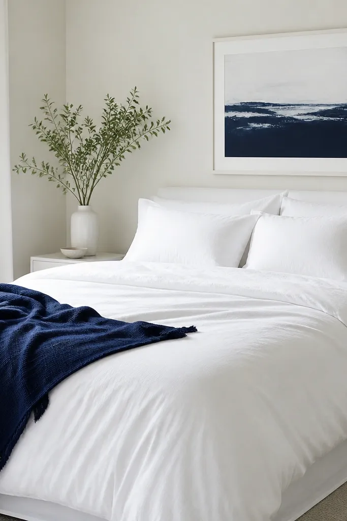 Bed is fully white: white duvet, white sheets, white pillows. One navy throw blanket is draped over the side. Above the bed is a navy-and-white framed print.