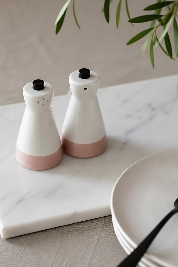 A white marble board on a table with a small salt and pepper set. The pieces are white with pink bases and tiny black tops. A white plate and black fork sit nearby.